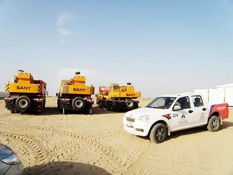 A white site pickup parked on sand in front of a line of mobile cranes